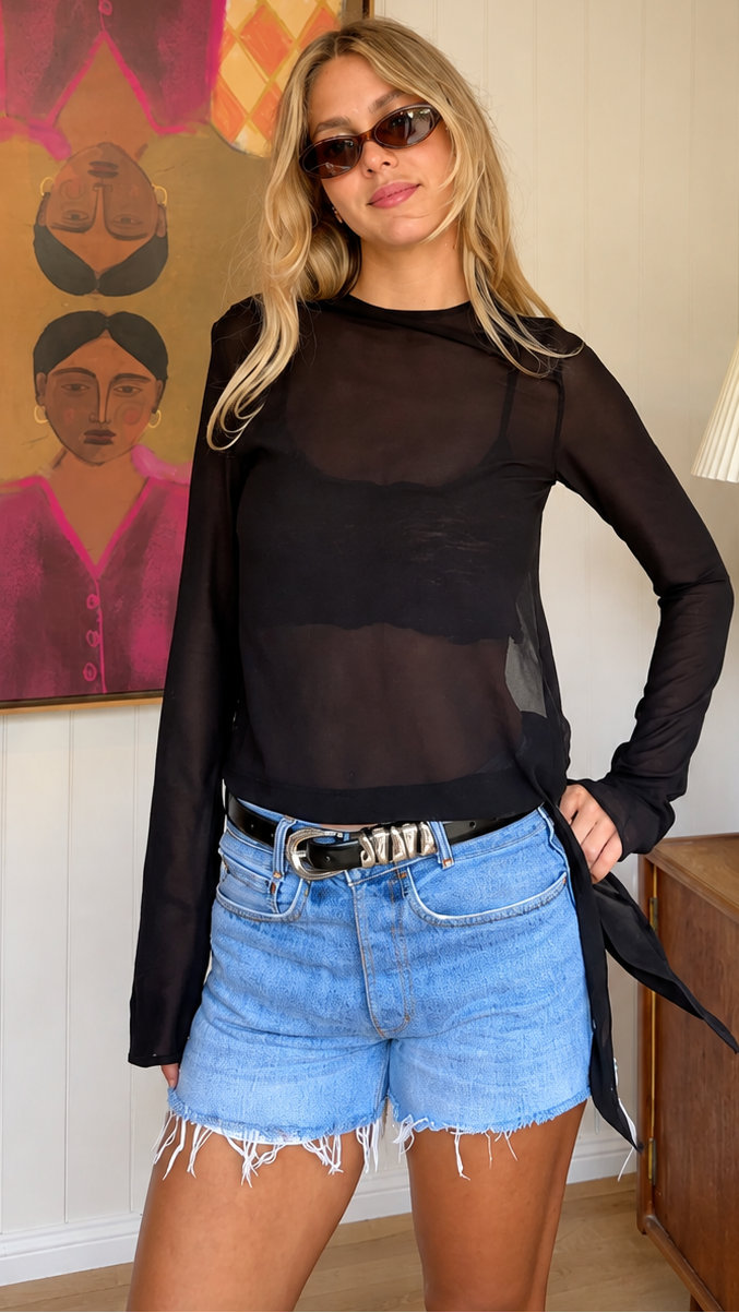 A woman modeling a sheer black long-sleeve mesh top with horizontal sheer bands, paired with denim shorts, in a bedroom setting.